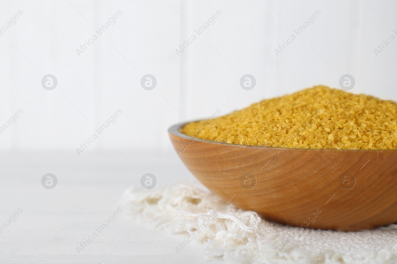 Photo of Napkin and bowl with uncooked bulgur on white wooden table, closeup. Space for text