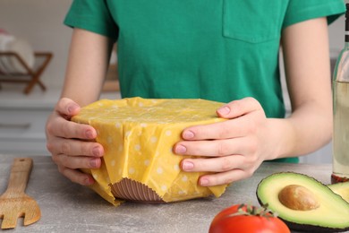 Photo of Woman holding bowl covered with beeswax food wrap at grey in kitchen, closeup