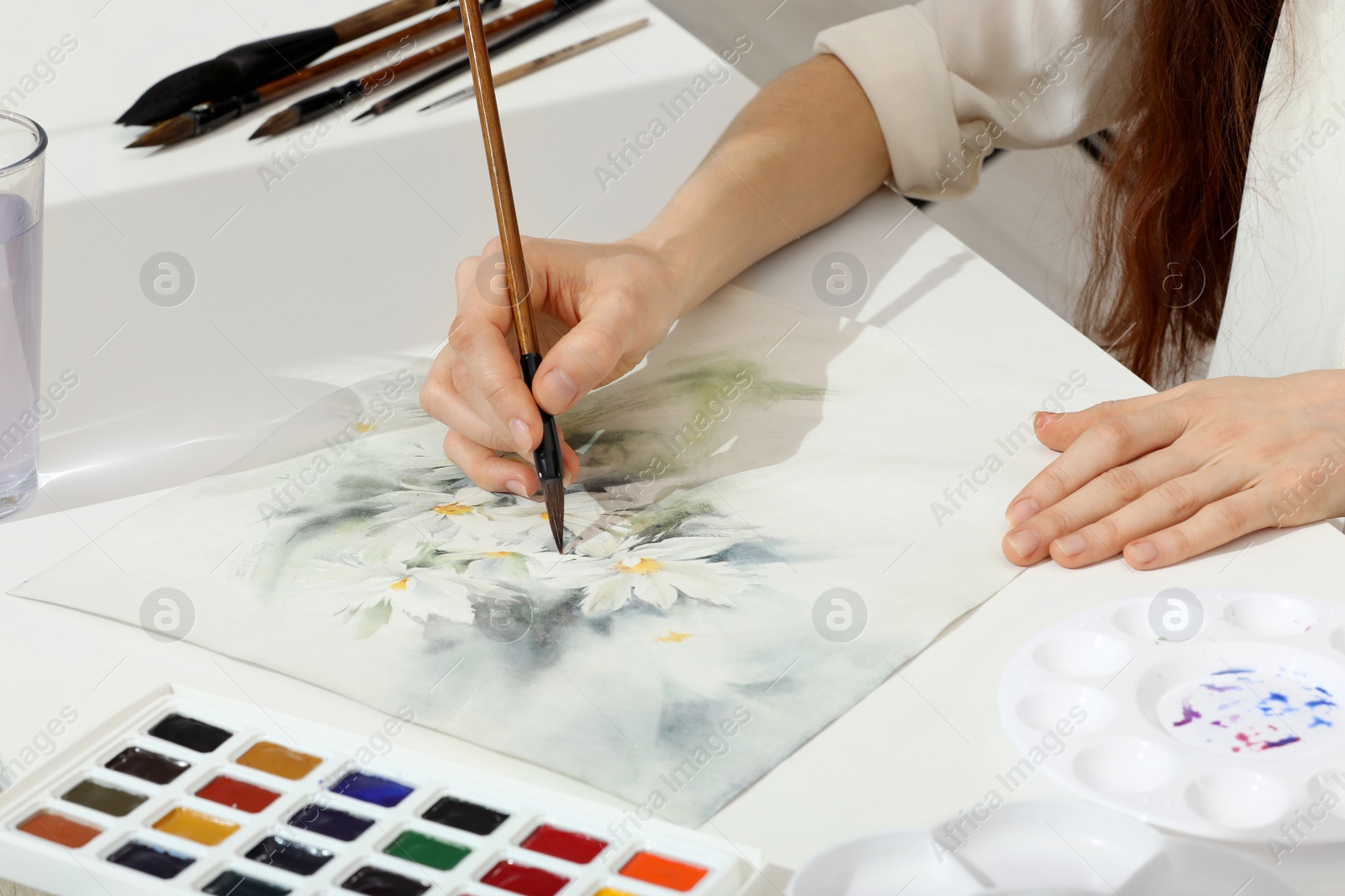 Photo of Woman painting flowers with watercolor at white table, closeup. Creative artwork