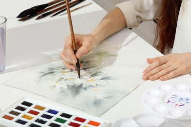 Photo of Woman painting flowers with watercolor at white table, closeup. Creative artwork
