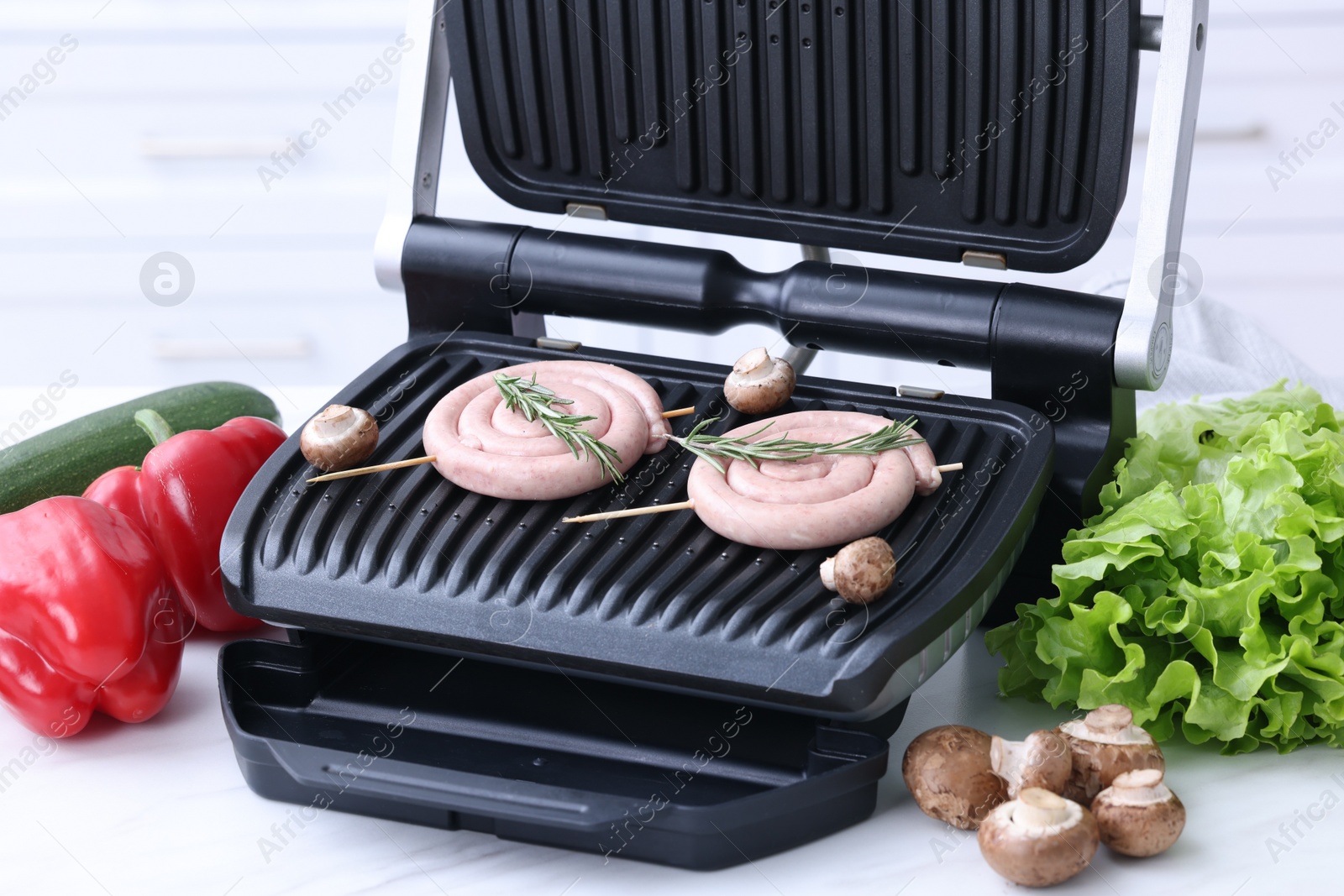 Photo of Electric grill with homemade sausages, rosemary and vegetables on white table indoors