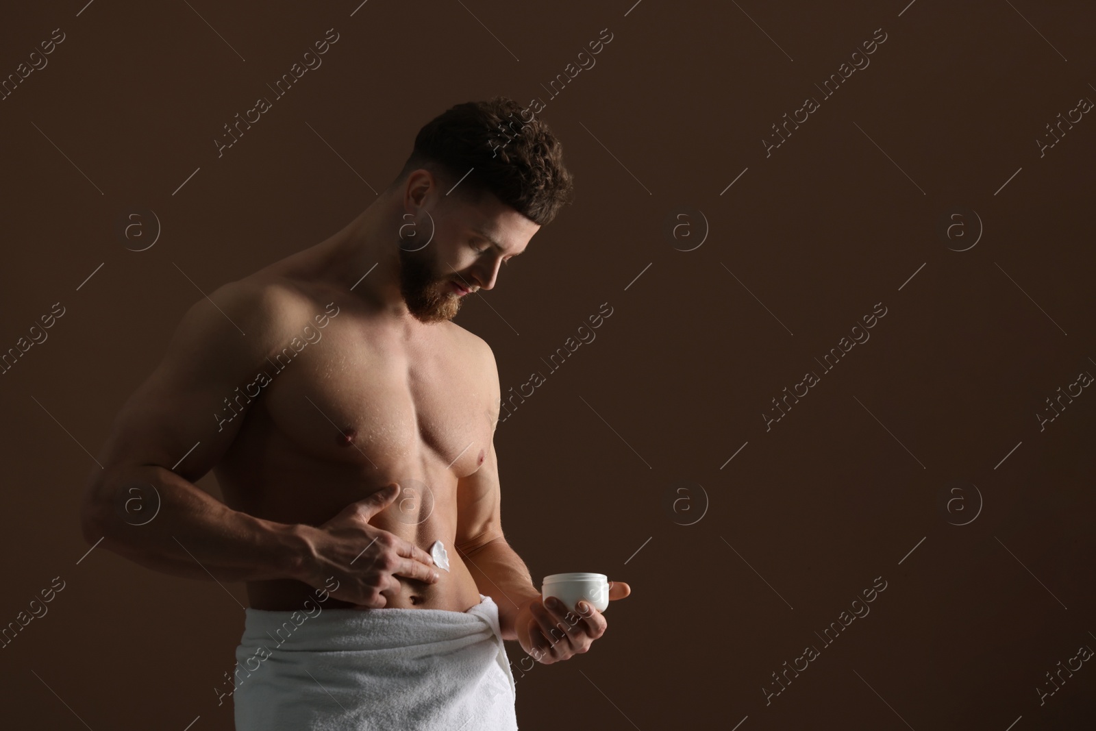 Photo of Handsome man applying moisturizing cream onto his body on brown background, space for text