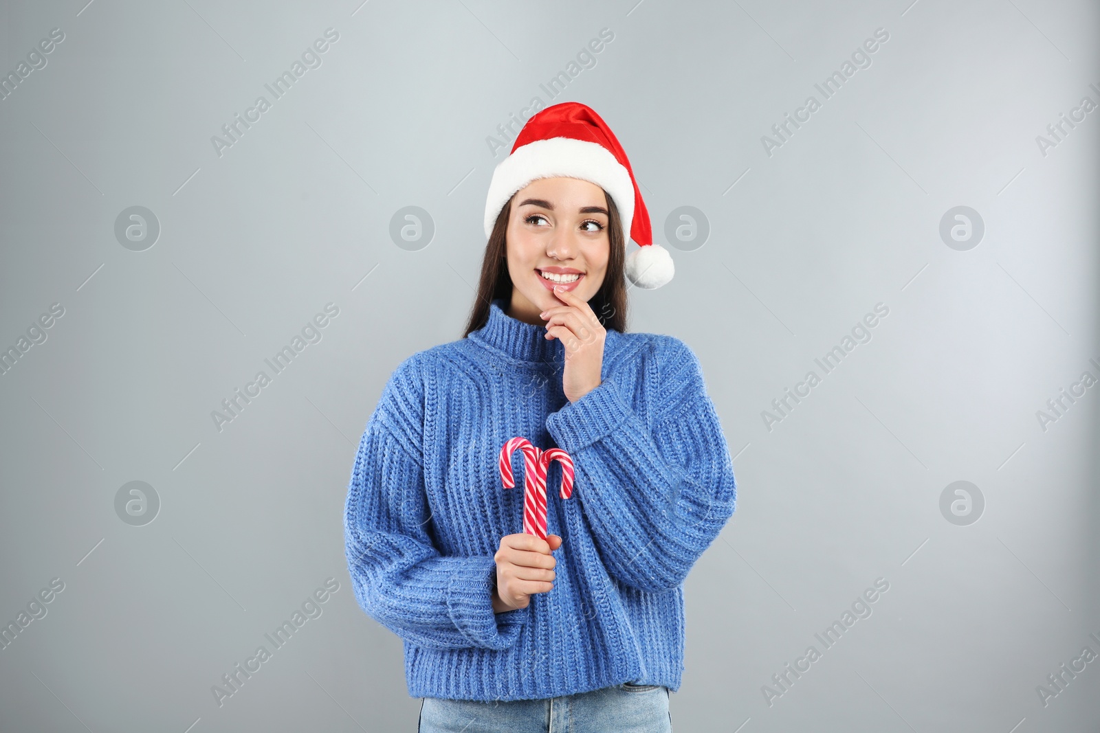 Photo of Young woman in blue sweater and Santa hat holding candy canes on grey background. Celebrating Christmas