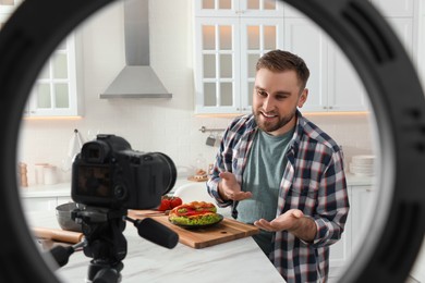 Blogger with tasty croissant sandwich recording video in kitchen at home, view through ring lamp