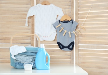 Photo of Basket with baby laundry, bottle of detergent and washing powder on white table