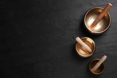 Photo of Golden singing bowls and mallets on black table, flat lay. Space for text
