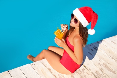 Photo of Young woman in Santa Claus hat with refreshing drink near swimming pool. Christmas vacation