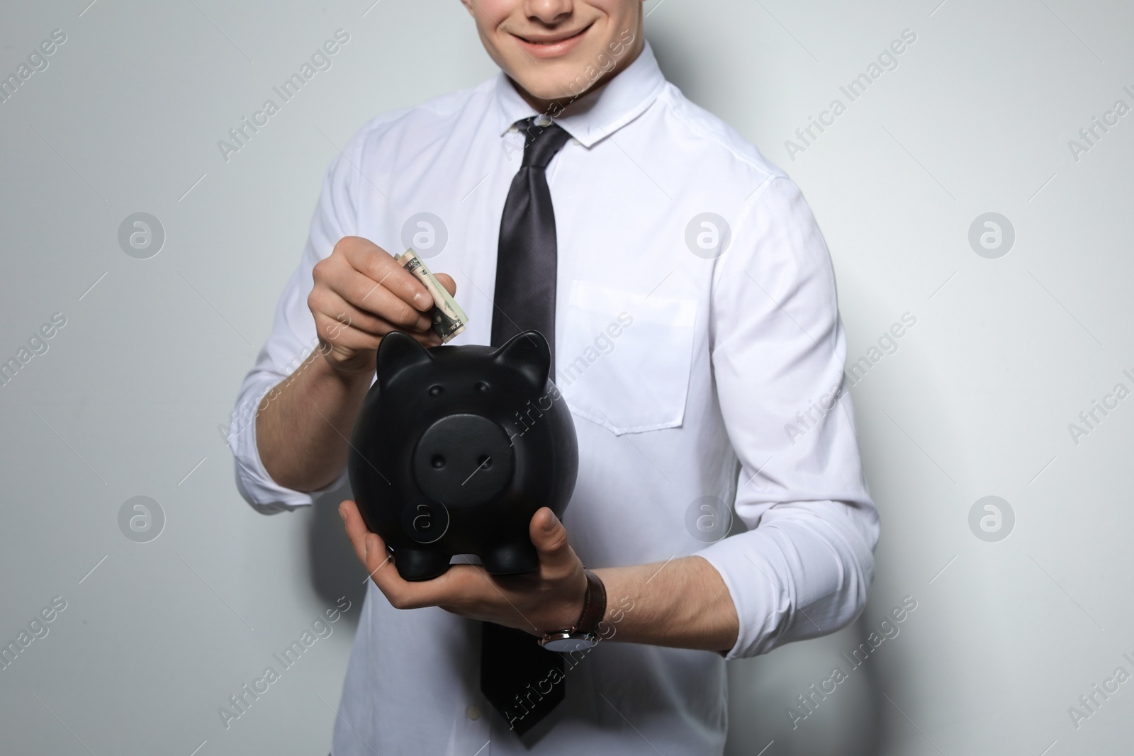 Photo of Young businessman putting money into piggy bank on light background, closeup