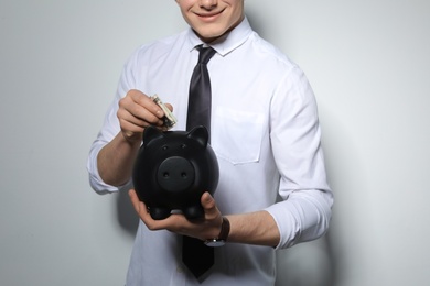 Photo of Young businessman putting money into piggy bank on light background, closeup