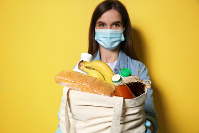 Photo of Female volunteer in protective mask and gloves with products against yellow background, focus on bag. Aid during coronavirus quarantine