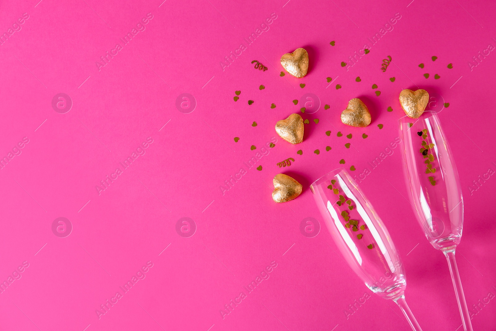 Photo of Champagne glasses and heart shaped candies in golden foil on color background, top view