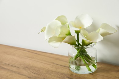 Beautiful calla lily flowers in glass vase on wooden table near white wall. Space for text