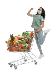 Young woman in medical mask with shopping cart full of groceries on white background