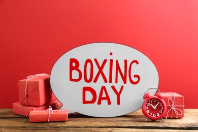 Composition with Boxing Day sign and Christmas gifts on wooden table against red background