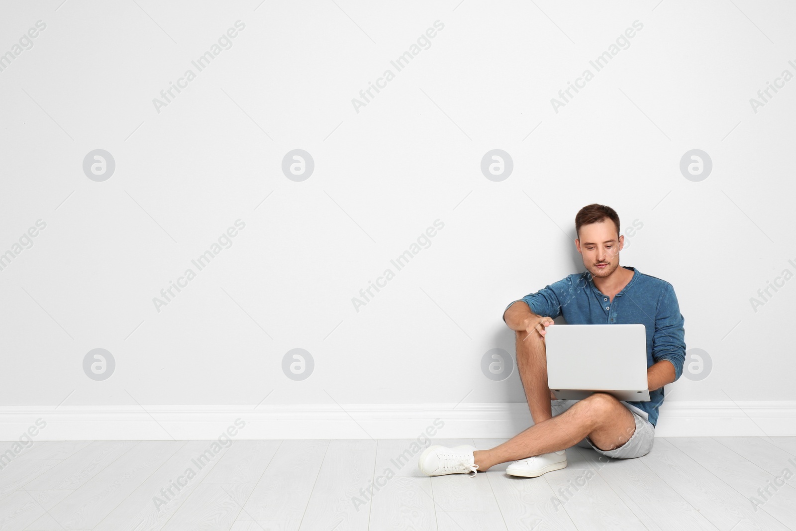 Photo of Young man with laptop sitting on floor against light wall. Space for text