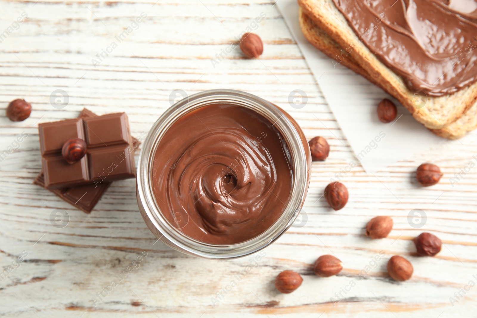 Photo of Flat lay composition with glass jar of tasty chocolate cream, bread and hazelnuts on wooden background