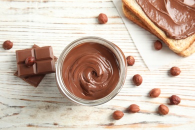 Photo of Flat lay composition with glass jar of tasty chocolate cream, bread and hazelnuts on wooden background