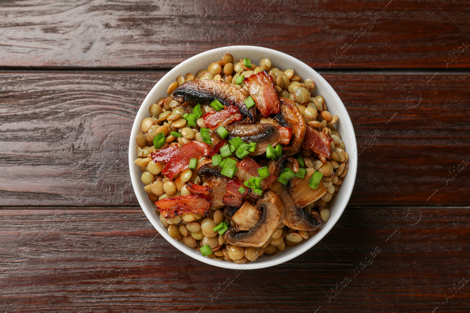 Photo of Delicious lentils with mushrooms, bacon and green onion in bowl on wooden table, top view