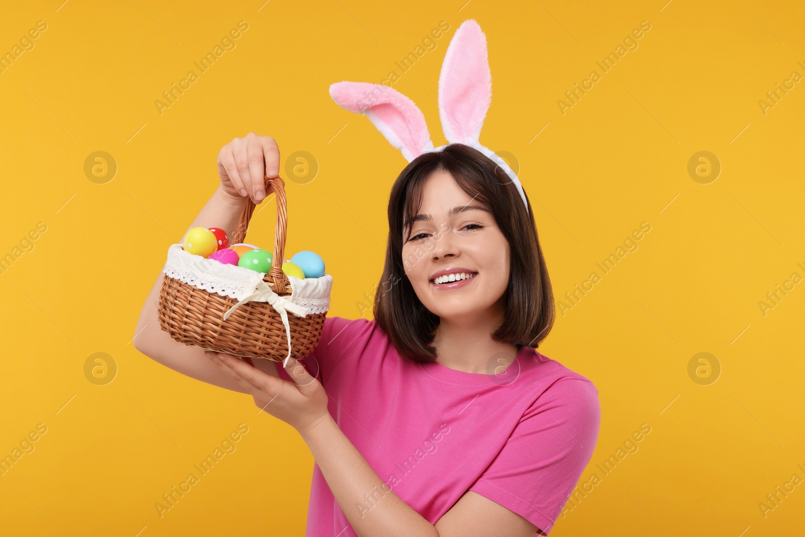 Photo of Easter celebration. Happy woman with bunny ears and wicker basket full of painted eggs on orange background