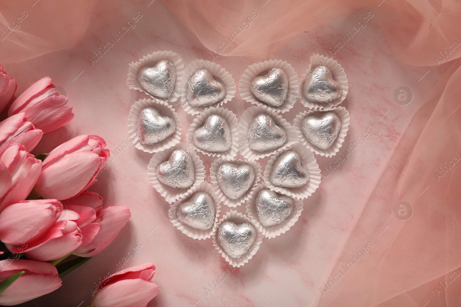 Photo of Heart made with delicious chocolate candies and beautiful tulips on pink table, flat lay