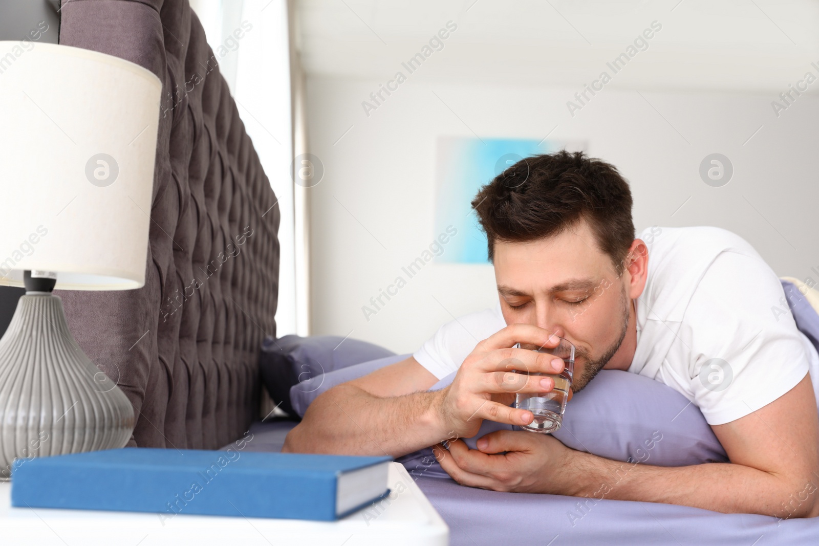 Photo of Handsome man drinking water while lying on pillow. Bedtime