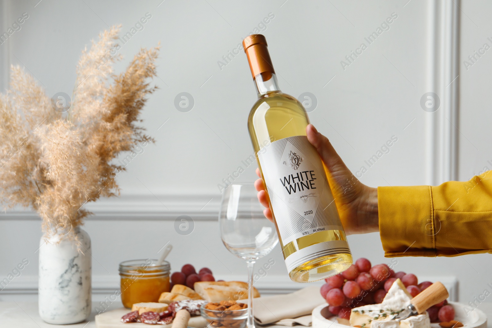 Photo of Woman holding bottle of white wine at table with snacks, closeup