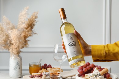 Photo of Woman holding bottle of white wine at table with snacks, closeup