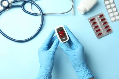 Photo of Doctor in latex gloves holding fingertip pulse oximeter near stethoscope and pills on light blue background, top view