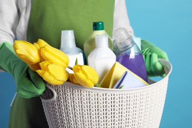 Photo of Spring cleaning. Woman holding basket with detergents, flowers and rags on light blue background, closeup