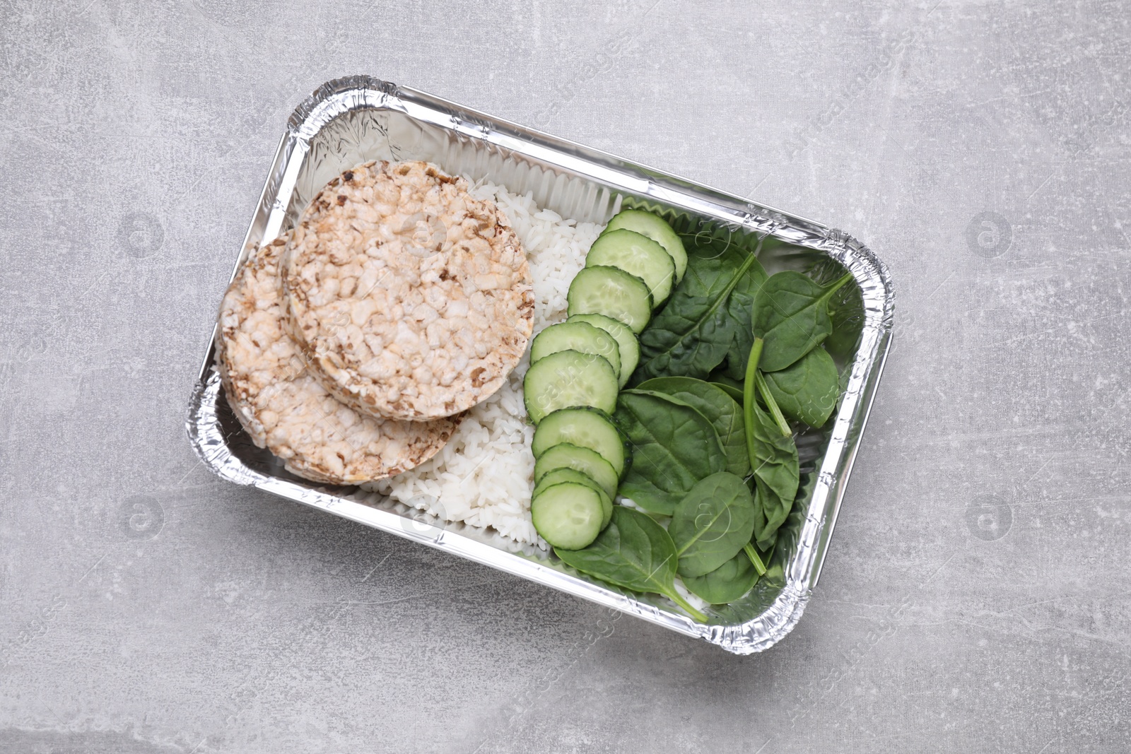 Photo of Container with rice, puffed crunchy cakes. fresh spinach and cut cucumbers on light grey table, top view