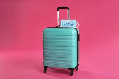 Photo of Stylish turquoise suitcase and protective masks with inscription COVID-19 on pink background. Travelling during coronavirus pandemic