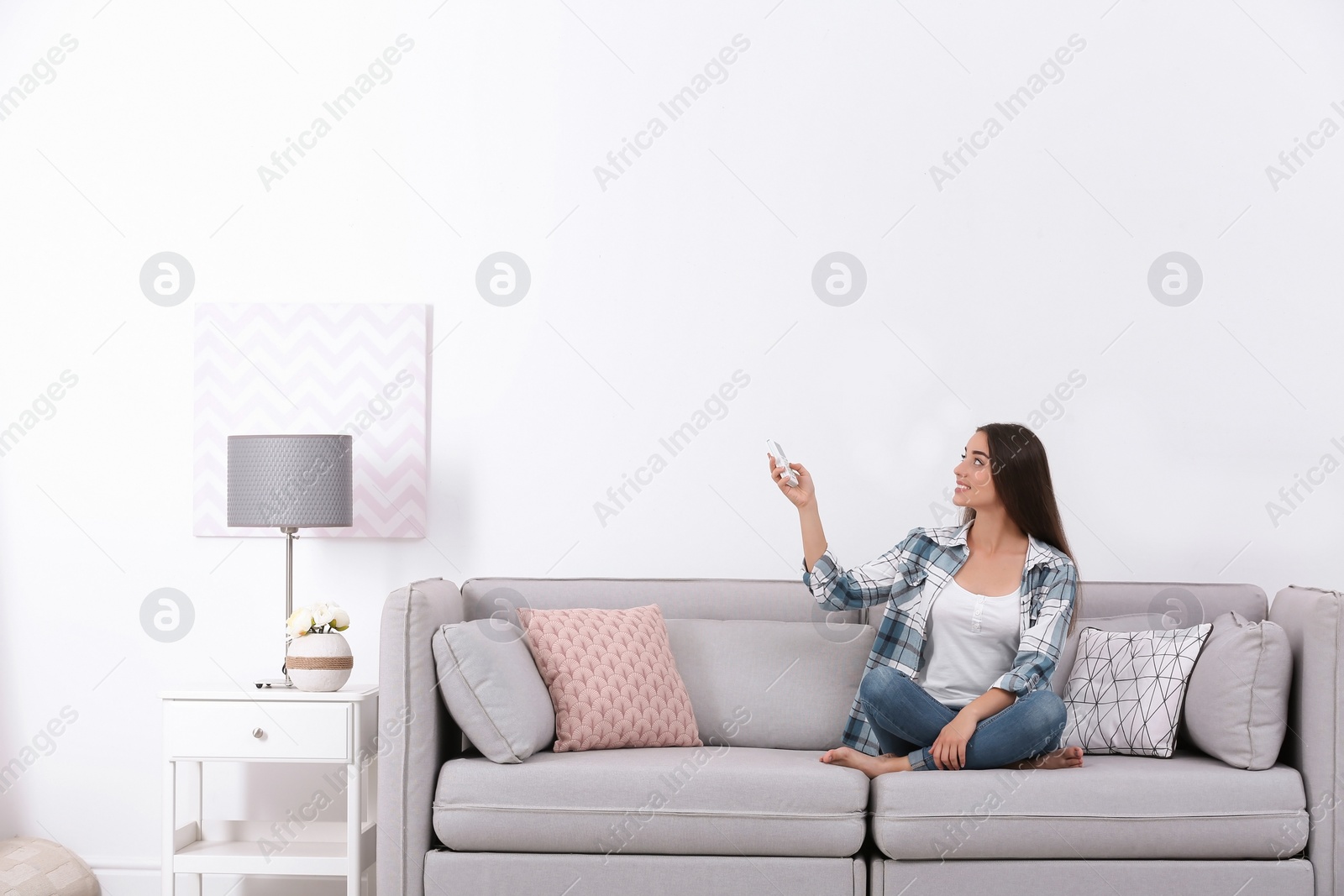 Photo of Young woman switching on air conditioner while sitting on sofa near white wall