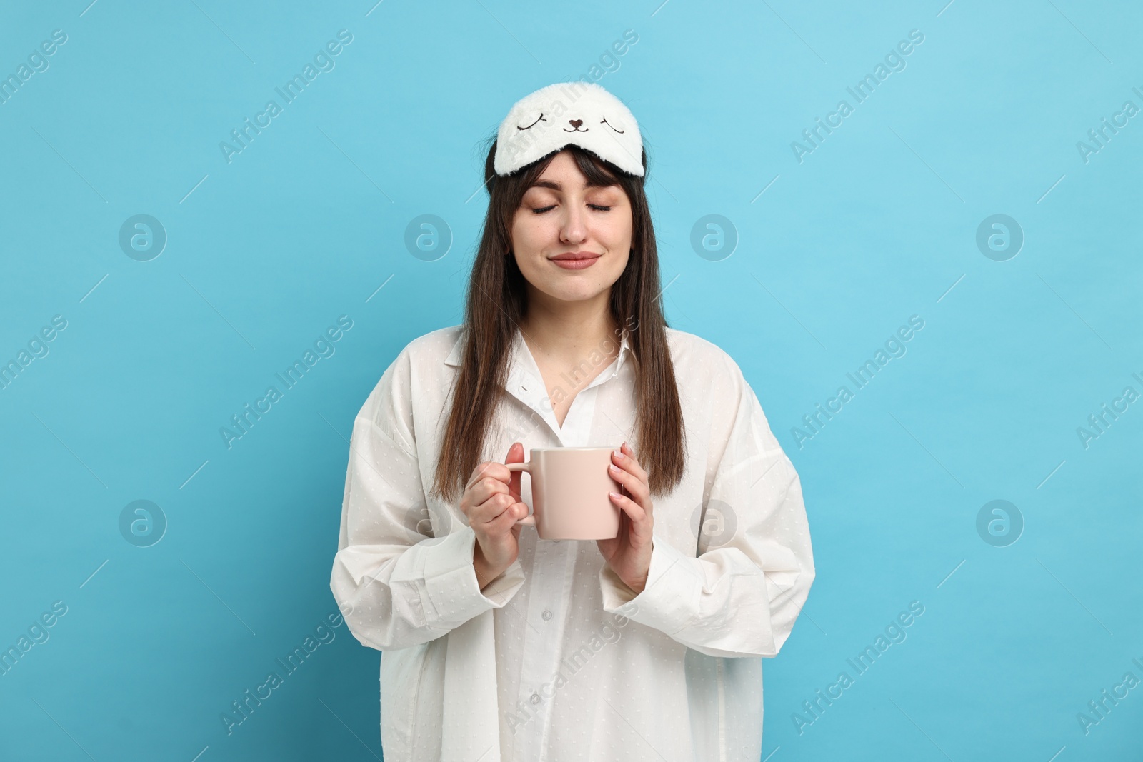 Photo of Woman in pyjama and sleep mask holding cup of drink on light blue background