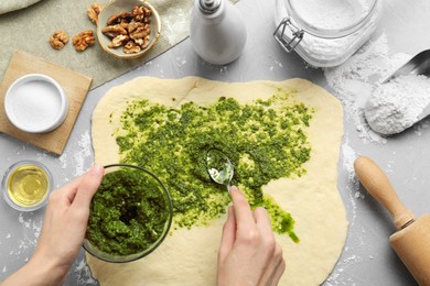 Photo of Making delicious pesto bread. Woman putting sauce onto raw dough at grey table, top view