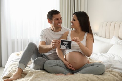 Young pregnant woman and her husband with ultrasound picture of baby in bedroom