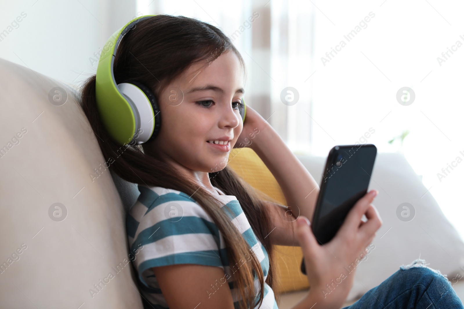 Photo of Cute little girl with headphones and smartphone listening to audiobook at home