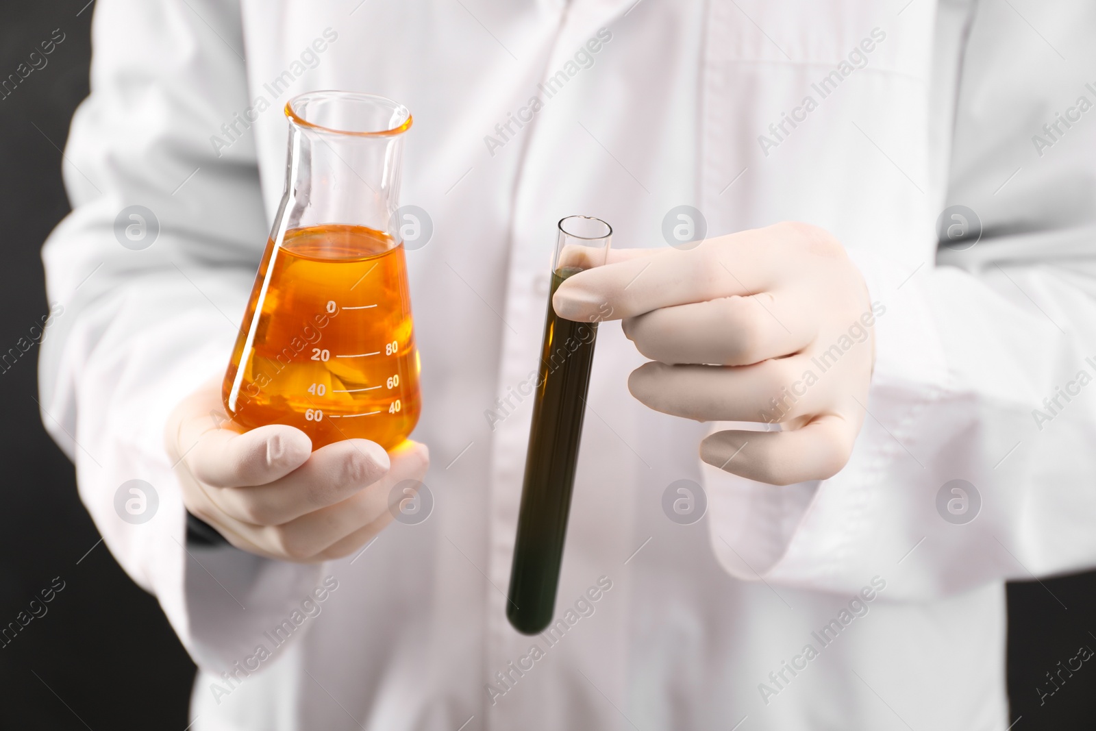 Photo of Woman holding test tube and flask with different types of crude oil on dark background, closeup