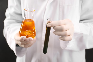 Photo of Woman holding test tube and flask with different types of crude oil on dark background, closeup