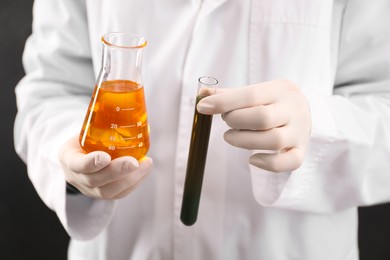 Woman holding test tube and flask with different types of crude oil on dark background, closeup