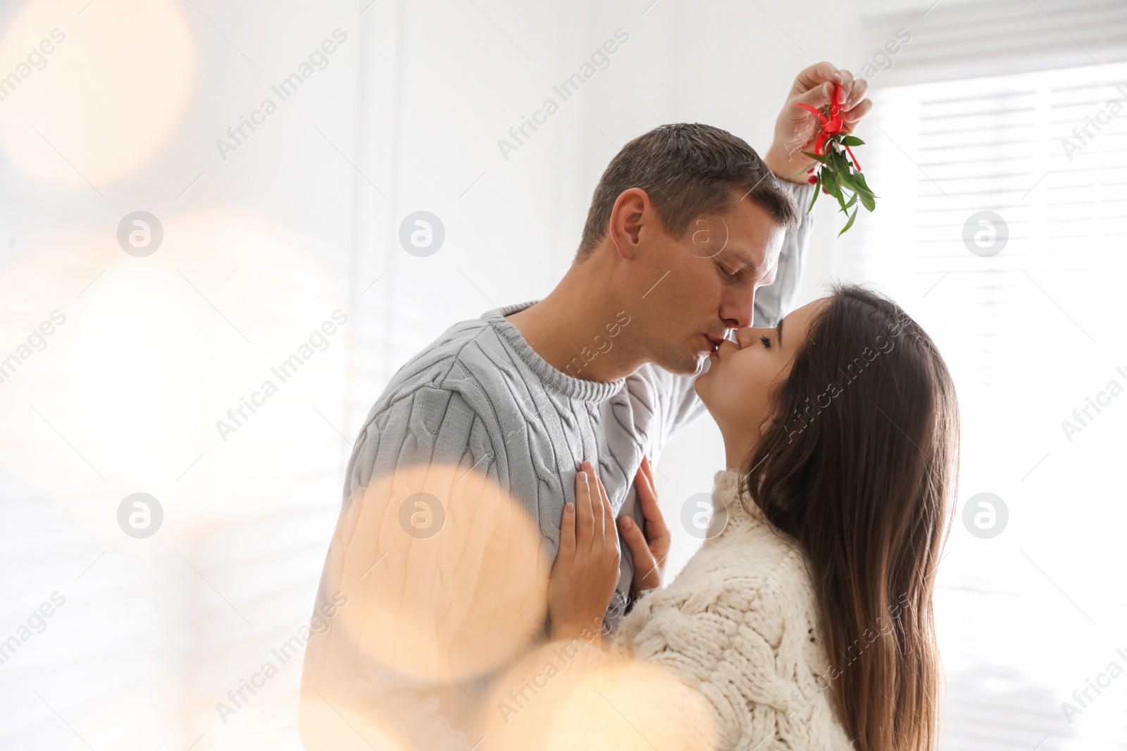 Photo of Happy couple kissing under mistletoe bunch at home, bokeh effect