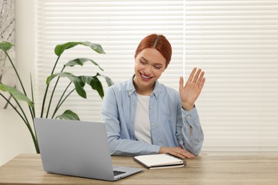 Photo of Young woman waving hello during video chat via laptop at table in office