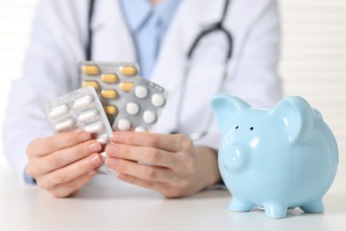 Doctor with pills and piggy bank at white table indoors, closeup