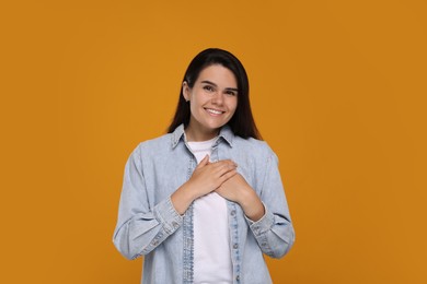 Photo of Thank you gesture. Beautiful grateful woman holding hands near heart on orange background