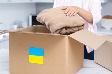 Photo of Humanitarian aid for Ukraine. Woman putting clothes in donation box at white table indoors, closeup