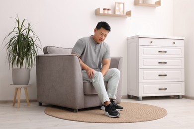 Photo of Asian man suffering from knee pain on armchair indoors