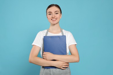 Beautiful young woman in clean apron with clipboard on light blue background