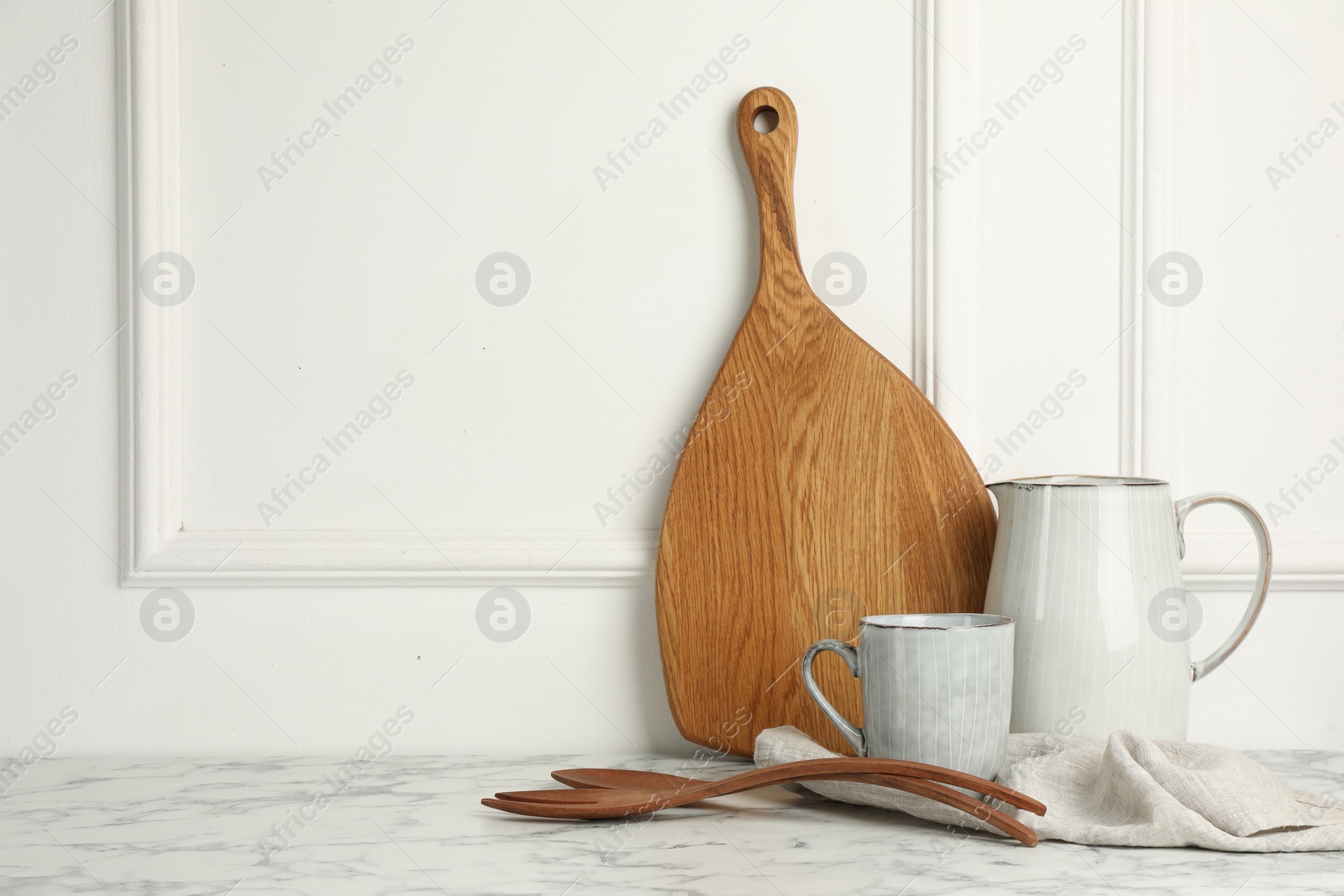 Photo of Wooden cutting board, kitchen utensils and dishware on white marble table. Space for text