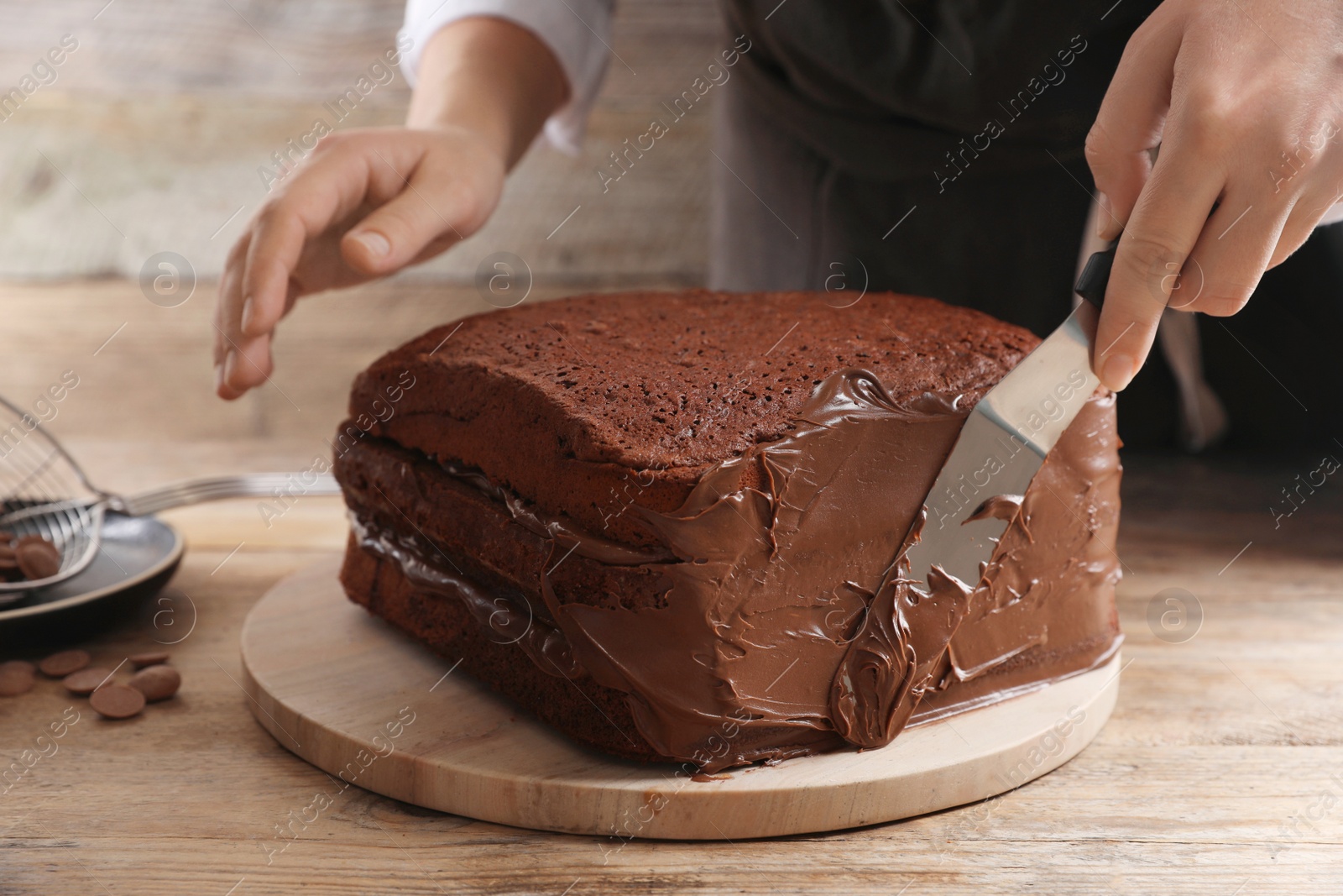 Photo of Woman smearing sides of sponge cake with chocolate cream at wooden table, closeup