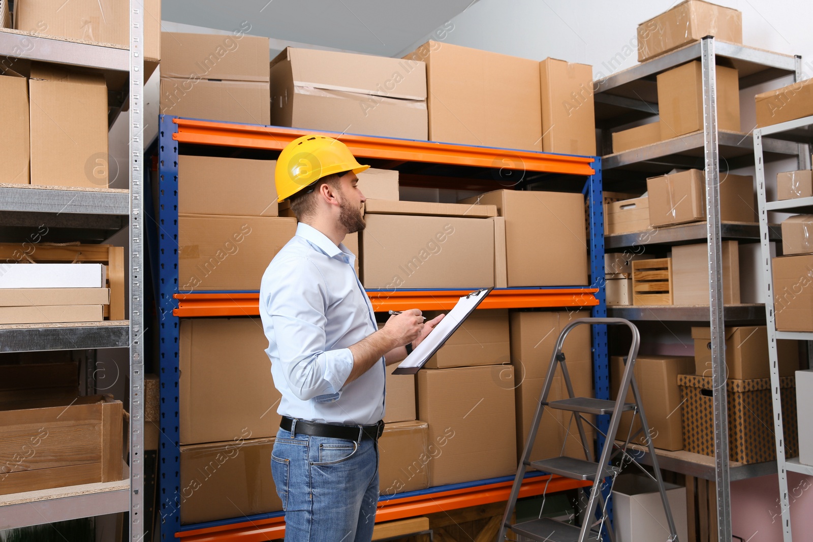 Photo of Young man with clipboard near rack of cardboard boxes at warehouse