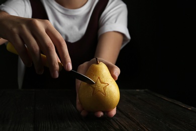 Photo of Woman carving star on ripe pear at table against dark background. Space for text